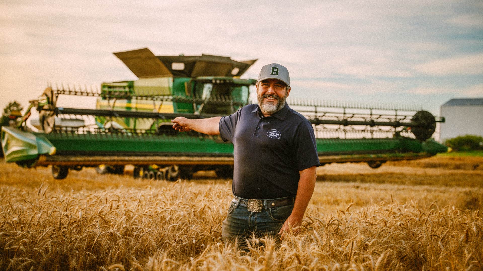 A farmer stands in a ripe field in front of a green combine.